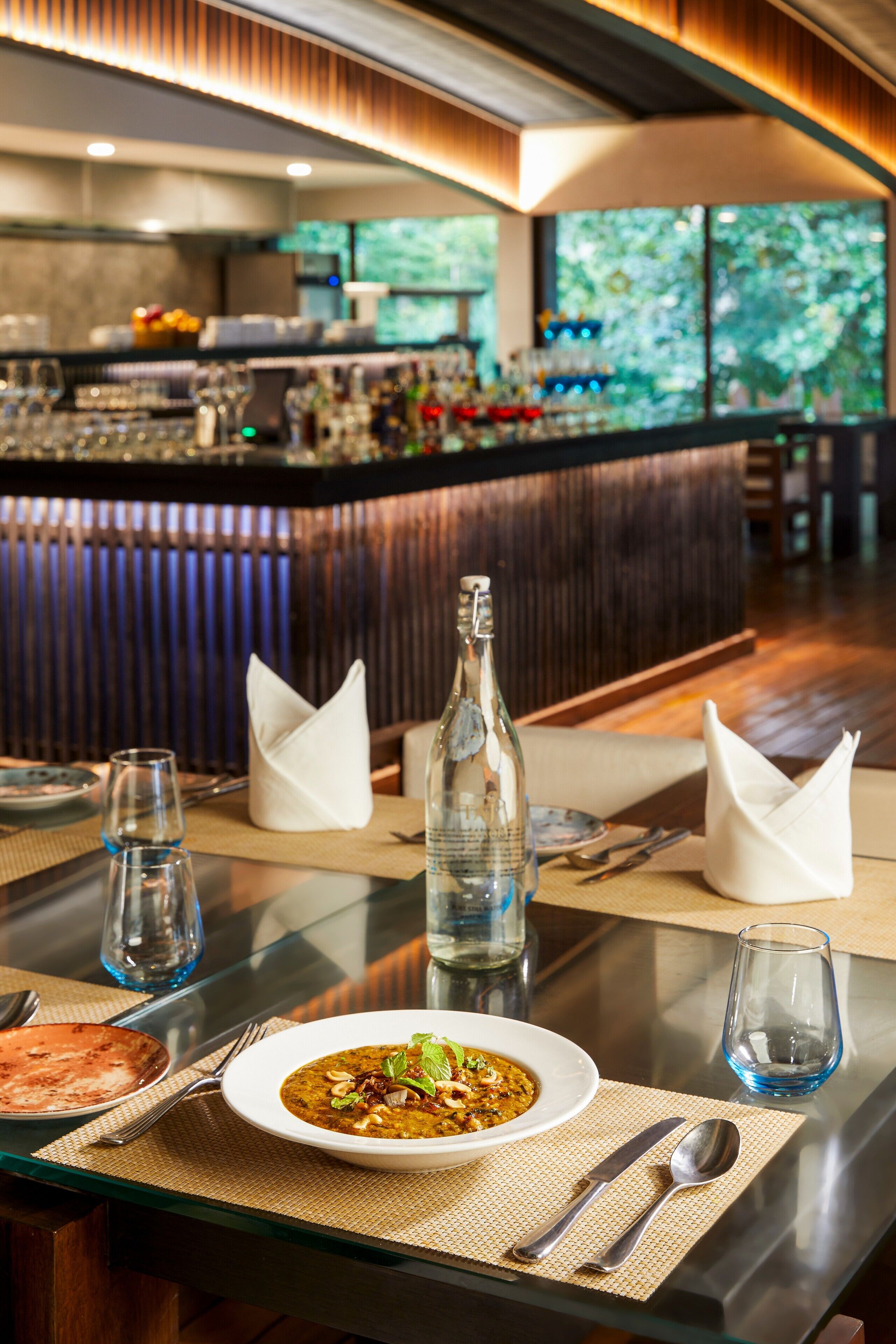 Une salle de restaurant avec une table dressée pour le dîner, un bar en arrière-plan et une vue sur la verdure à l’extérieur.