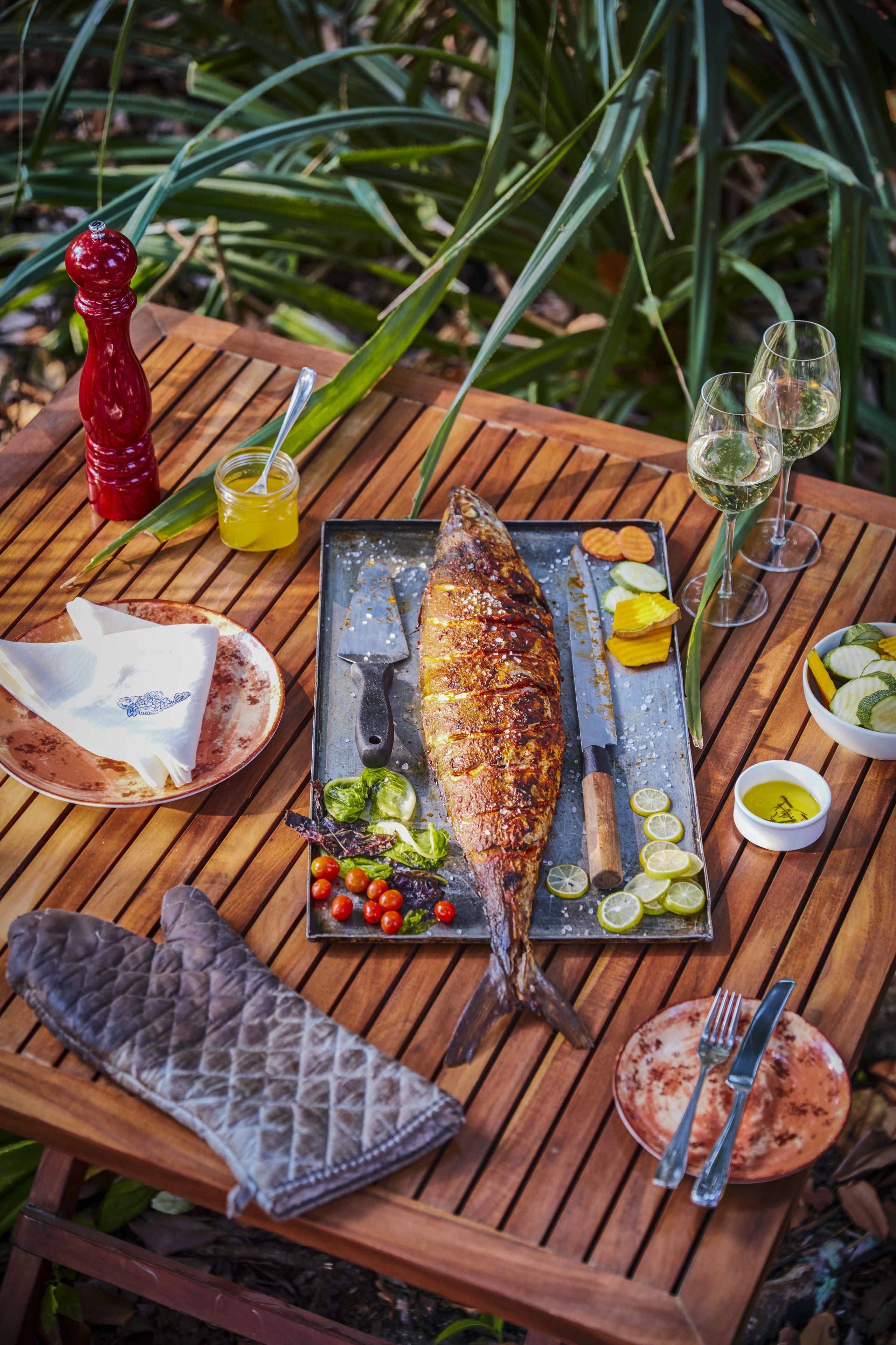 Una mesa de madera con un pescado asado, cubiertos, un cuchillo, un tenedor, un molinillo de pimiento rojo, una copa de vino blanco y una copa de vino rosado.