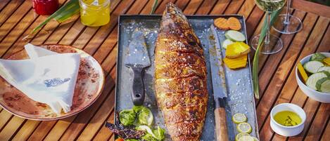 Una mesa de madera con un pescado asado, cubiertos, un cuchillo, un tenedor, un molinillo de pimiento rojo, una copa de vino blanco y una copa de vino rosado.