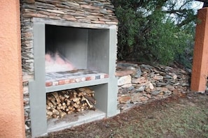 An outdoor stone fireplace with a fire burning inside, surrounded by a stone wall and a pile of firewood. - Matjiesvlei Retreat 1945 (Calitzdorp)
