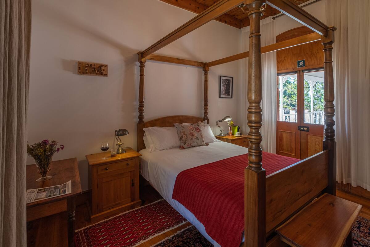 A four-poster bed with a wooden frame, a red throw blanket, and a nightstand with a lamp and a vase of flowers.