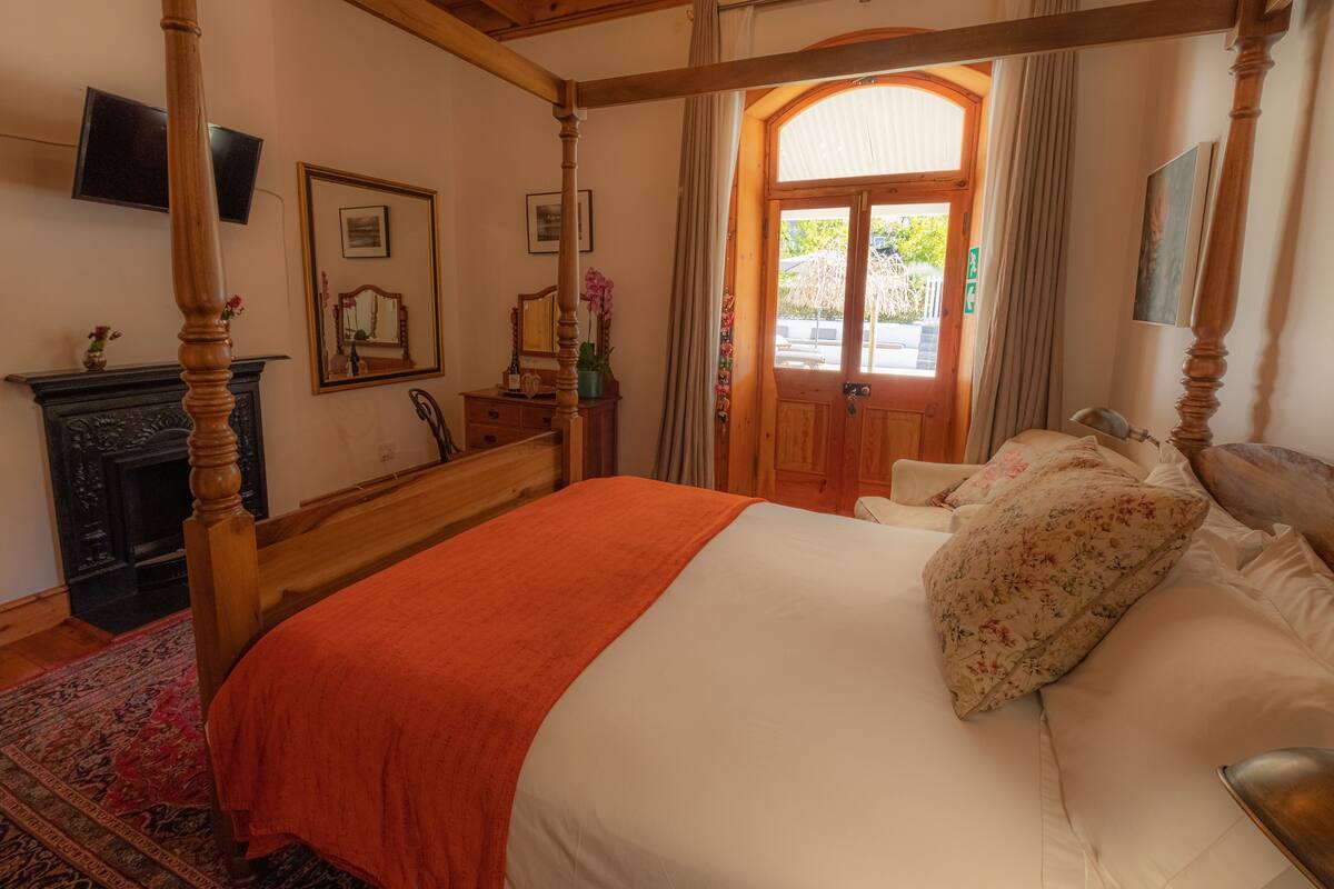 A four-poster bed with a red bedspread, a wooden headboard, and a floral pillow. There is a wooden dresser with a mirror and a vase of flowers. A fireplace is visible in the corner, and a television is mounted on the wall.