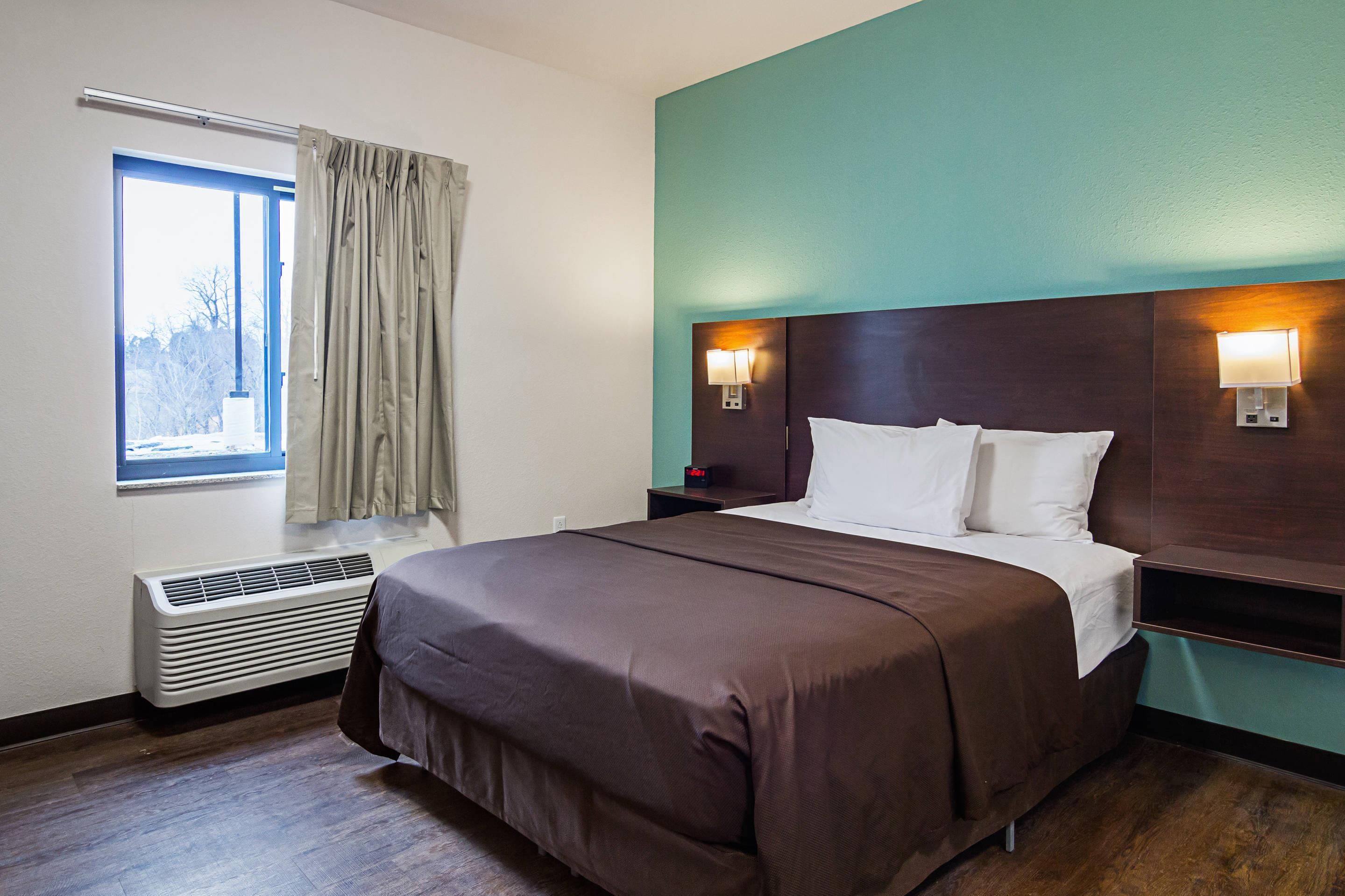 A hotel room with a bed, a window with curtains, a wall-mounted heater, and two wall sconces.