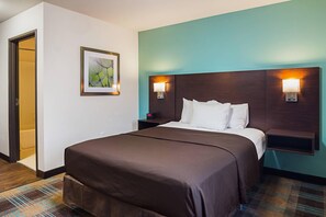 A hotel room with a bed, a nightstand, a wall-mounted lamp, and a door leading to a bathroom. - Suburban Studios Monaca - Pittsburgh (Monaca)