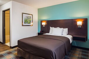 Habitación de hotel con cama, mesita de noche, lámpara de pared y puerta que da a un baño.