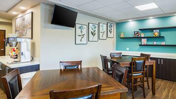 A restaurant with a wooden dining table, chairs, a flat-screen TV mounted on the wall, framed botanical prints, and shelves with children's books and games.