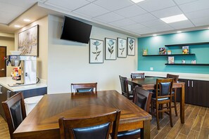 A restaurant with a wooden dining table, chairs, a flat-screen TV mounted on the wall, framed botanical prints, and shelves with children's books and games. - Suburban Studios Monaca - Pittsburgh (Monaca)