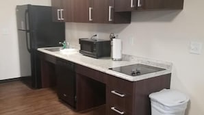 A compact kitchen with dark wooden cabinets, a black refrigerator, a microwave, and a trash bin. - Suburban Studios Monaca - Pittsburgh (Monaca)