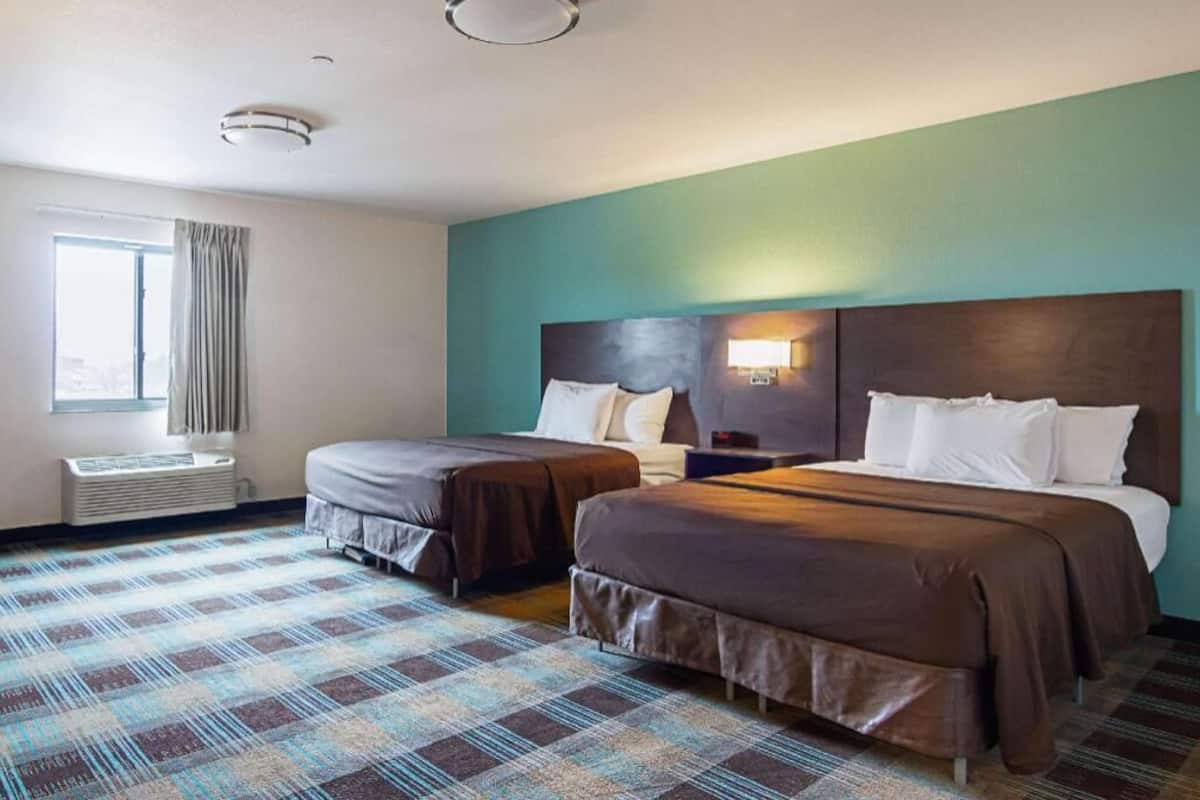 A hotel room with two beds, a window with curtains, a wall-mounted heater, and a check-in desk.