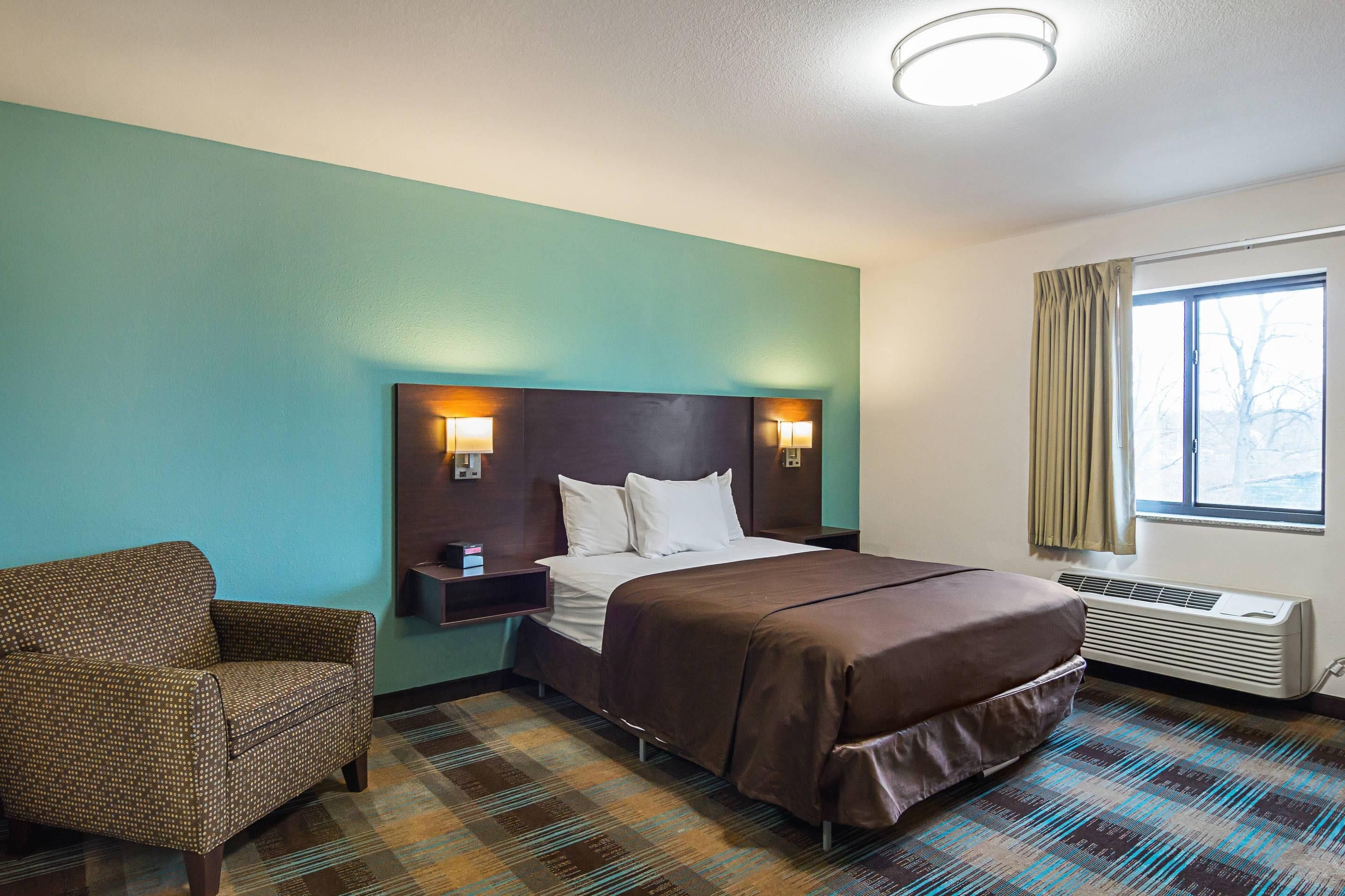 A hotel room with a bed, nightstand, chair, and window.
