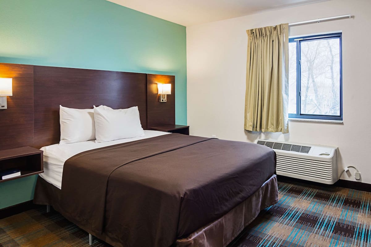 A hotel room with a large bed, a headboard with wall-mounted lights, a window with curtains, and a wall-mounted air conditioner.