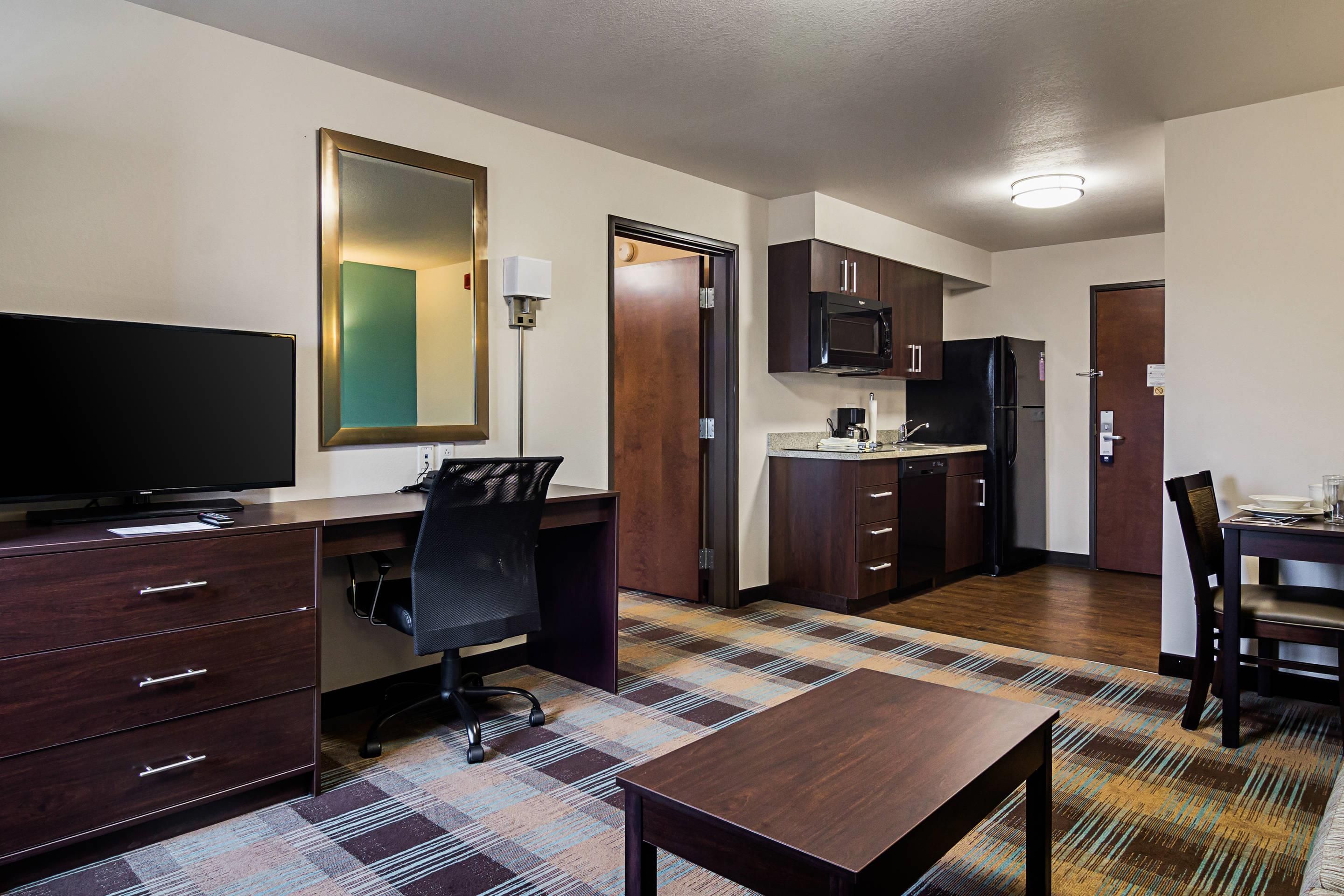 A compact hotel room with a desk, chair, television, kitchenette, and a patterned carpet.