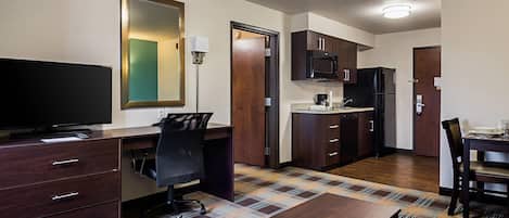 A compact hotel room with a desk, chair, television, kitchenette, and a patterned carpet.