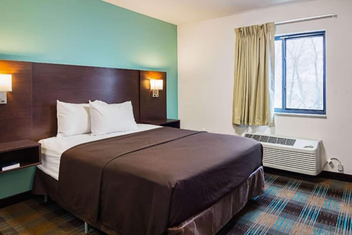 A hotel room with a large bed, a nightstand, a wall-mounted lamp, a window with curtains, and an air conditioning unit.