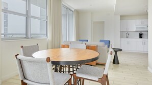 A modern, well-lit room with a round wooden table and grey upholstered chairs. Large windows with curtains allow natural light, and a kitchen area is visible in the background. - Circ Hotel (Hollywood)
