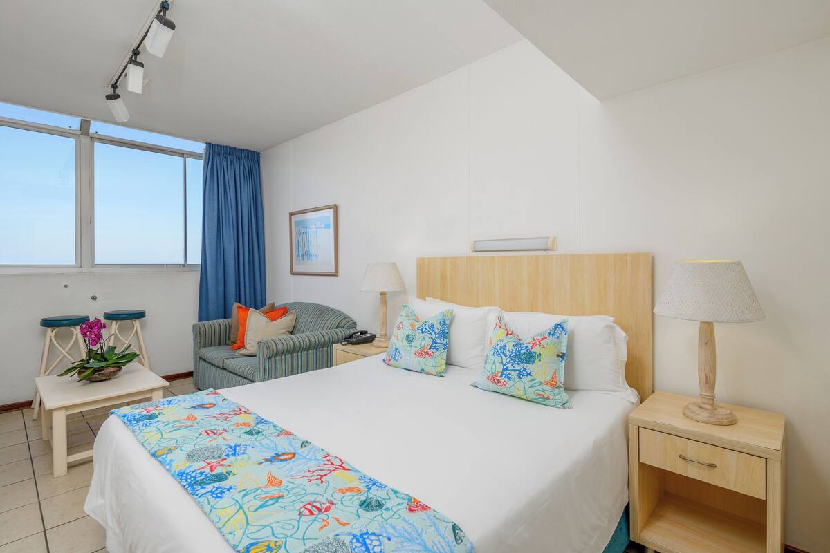 A hotel room with a large bed, two bedside tables, a chair, a small table with a plant, and a window with curtains.
