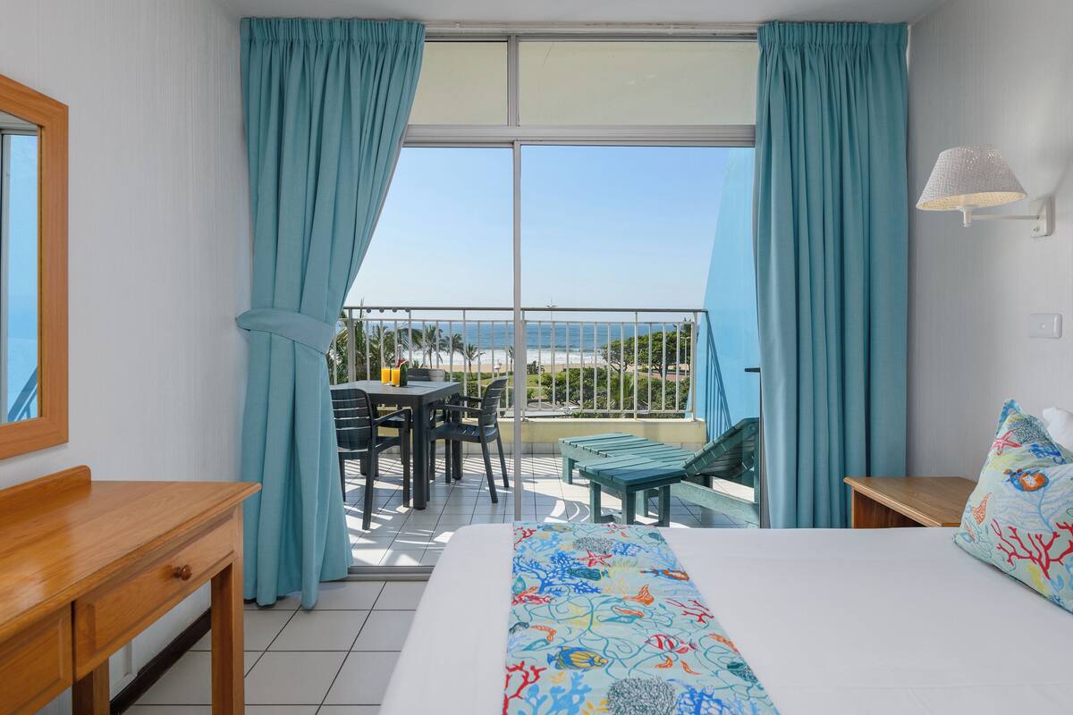A hotel room with a balcony, a bed with a colorful bedspread, a wooden nightstand, and a table with chairs outside.