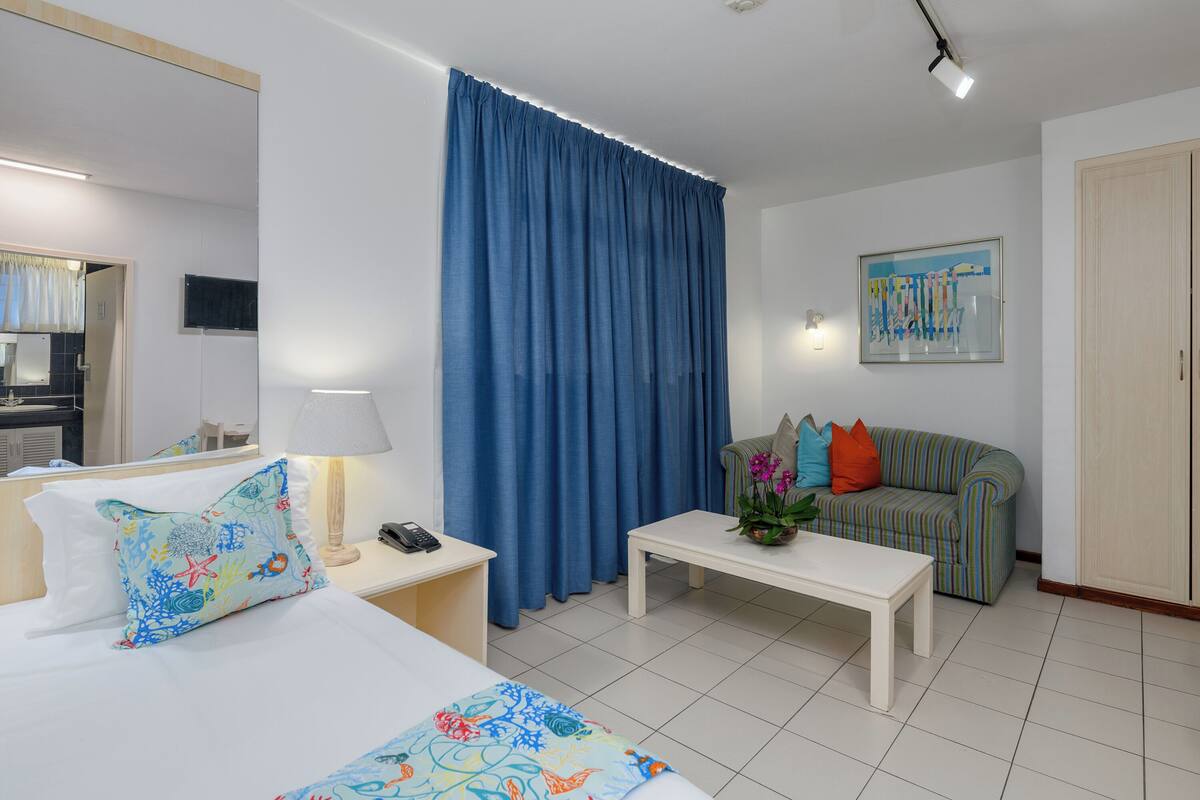A hotel room with a bed, a nightstand, a bedside table, a sofa, a coffee table, a curtain, a painting, and a door.