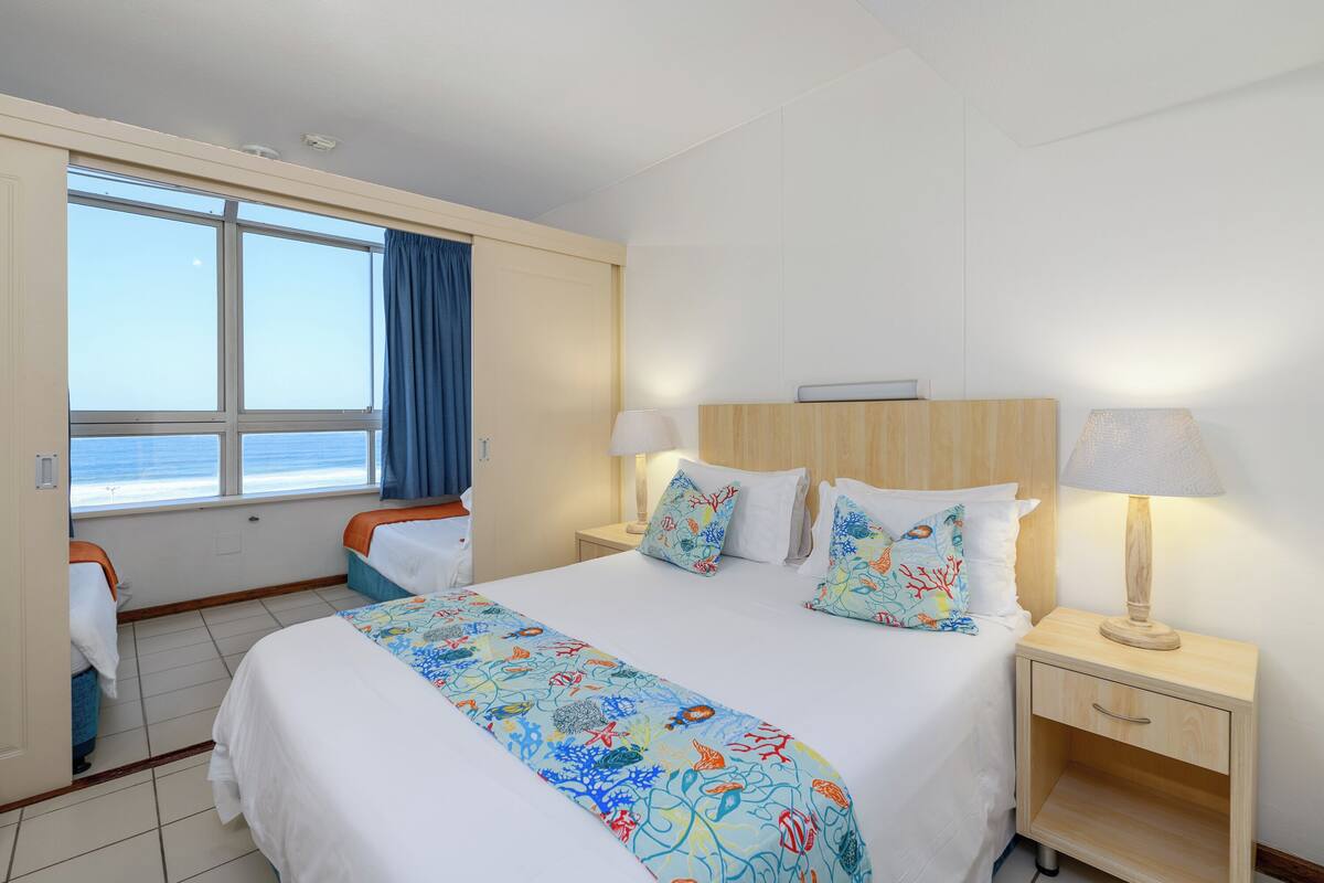 A hotel room with a large bed, two bedside tables, a window with a view of the sea, and a door leading to a balcony.