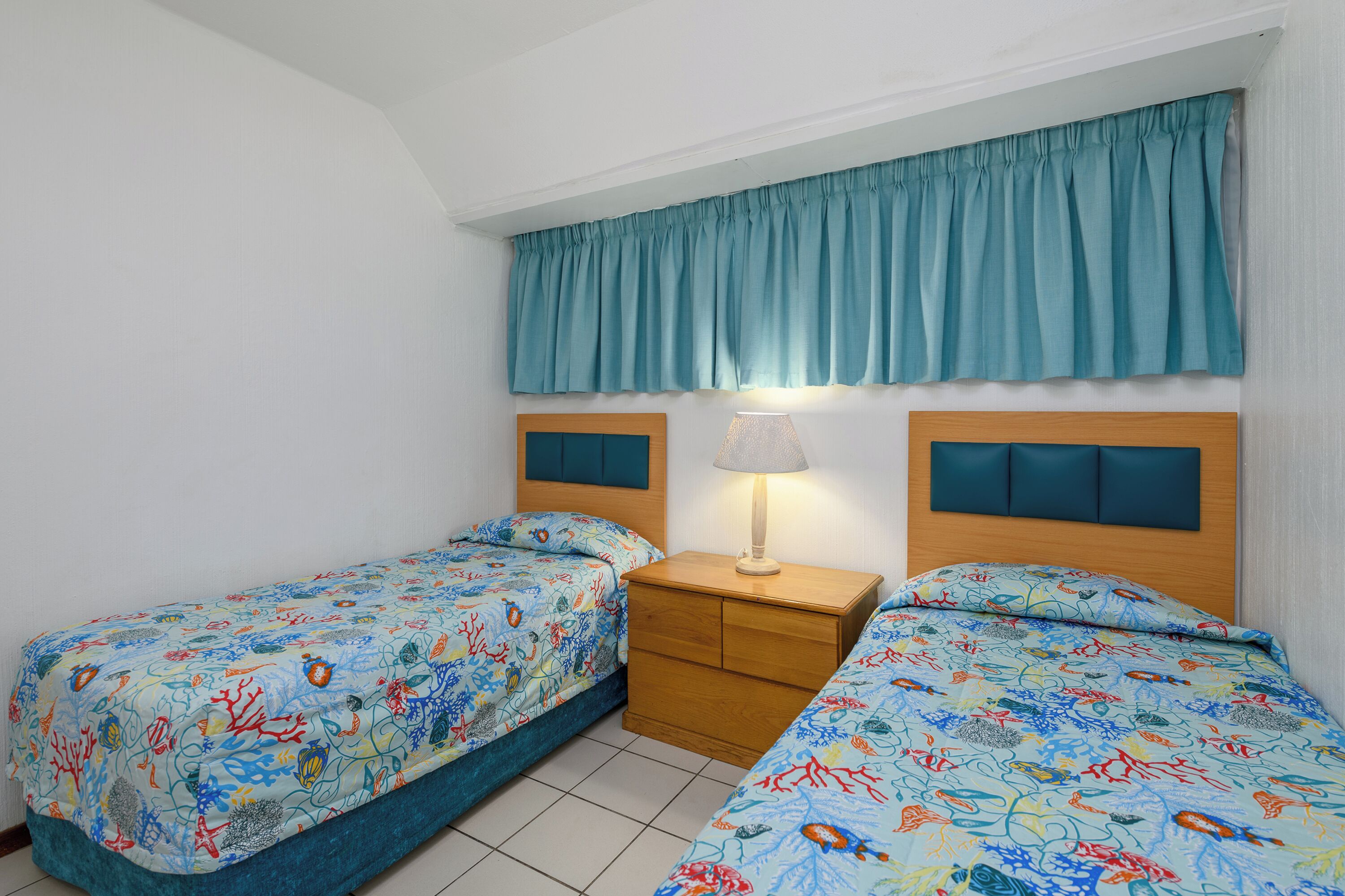 Un dormitorio con dos camas, una mesita de noche, una lámpara y una cortina color aguamarina.