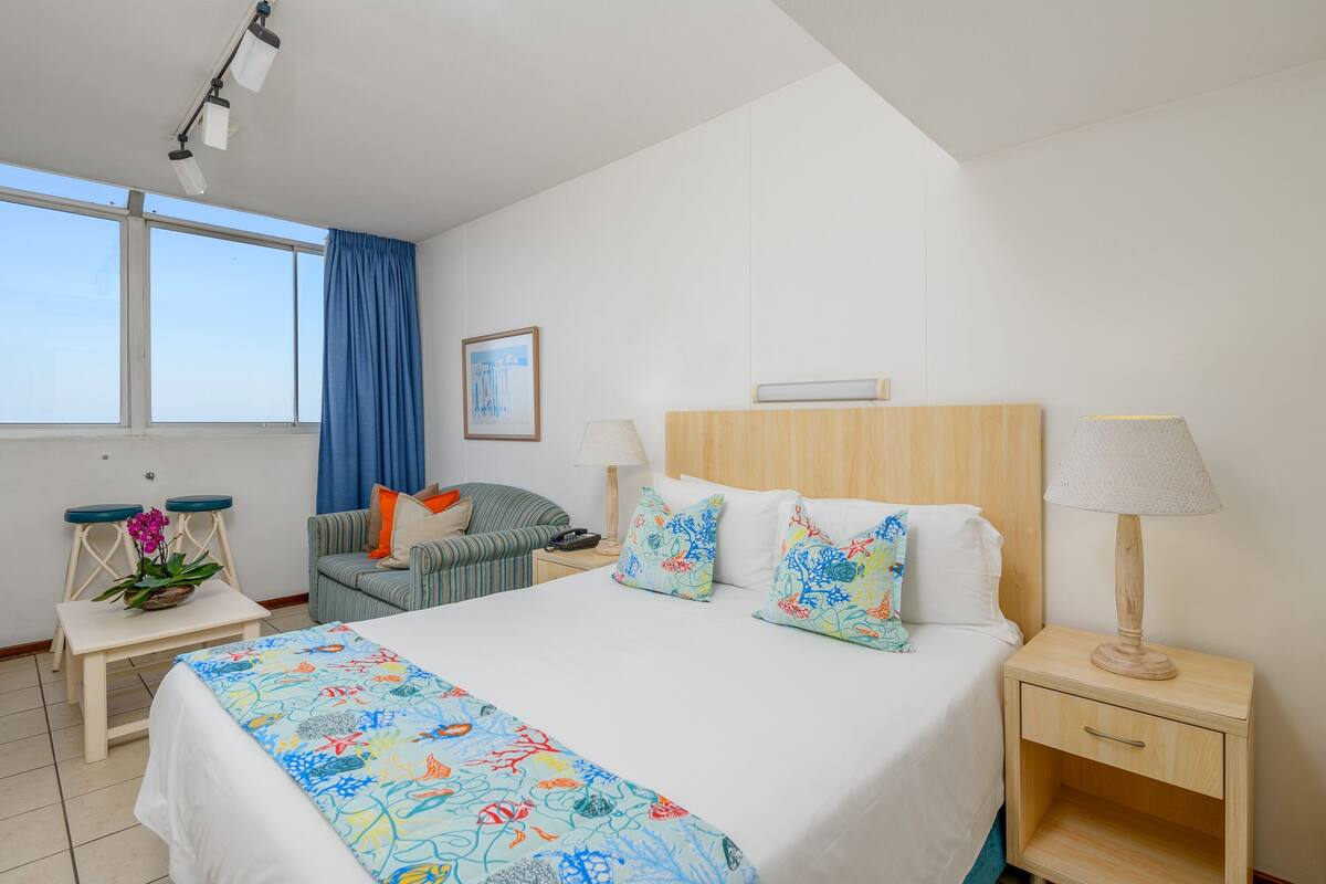 A hotel room with a bed, bedside tables, a chair, a desk with a plant, and a window with curtains.