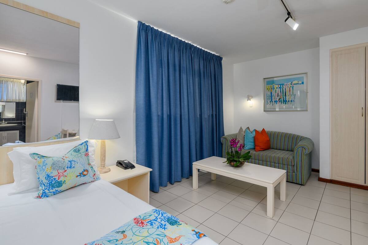 A hotel room with a bed, a bedside table with a lamp, a coffee table, a sofa with cushions, and a large window with blue curtains.