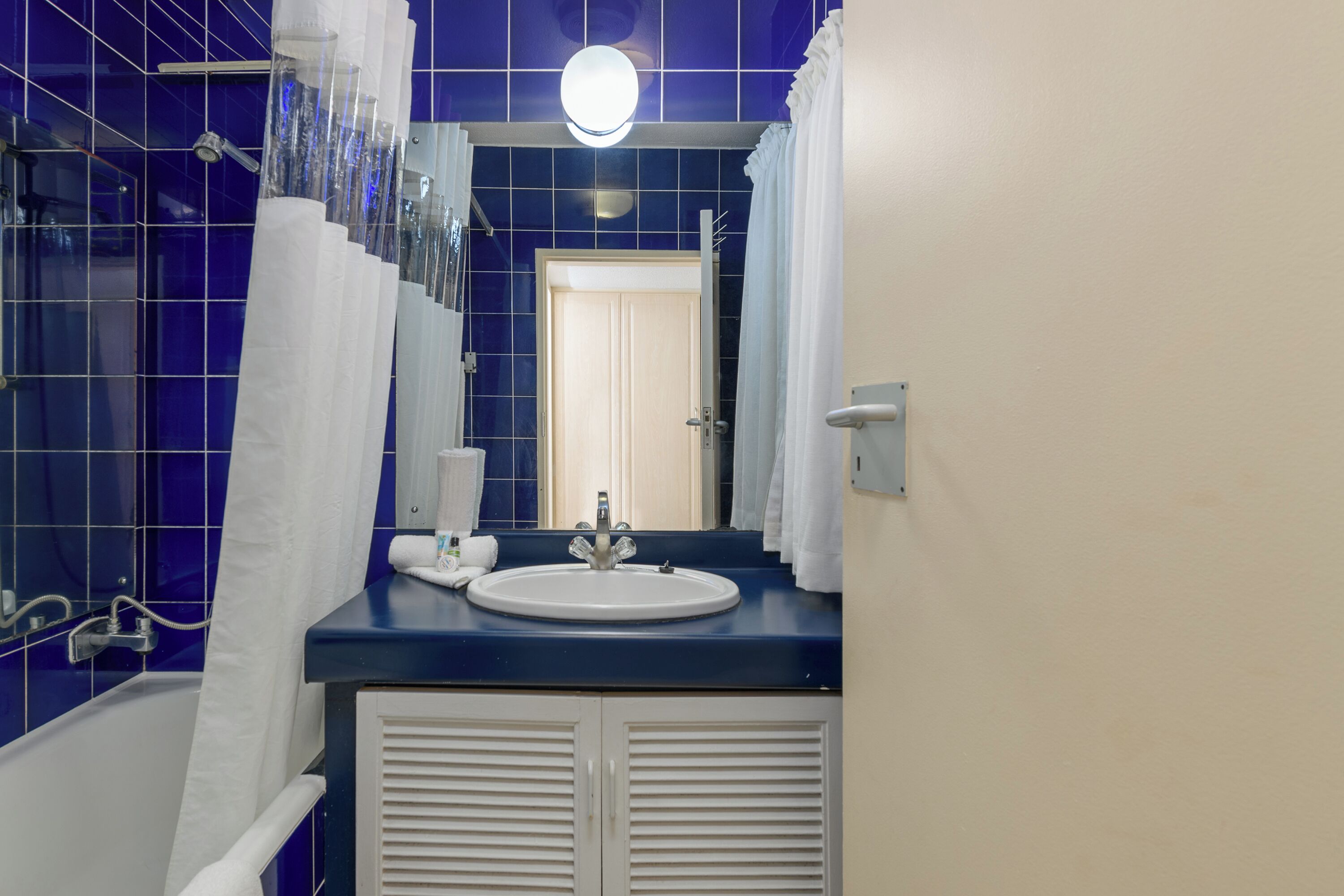 Een badkamer met een blauwe betegelde muur, een witte wastafel, een spiegel en een douche met een witte gordijn.