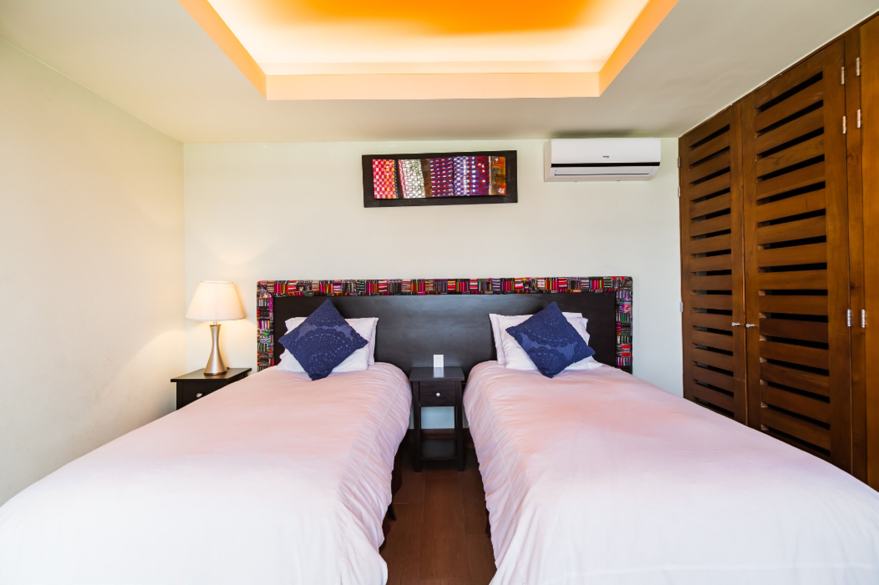 A hotel room with two beds, a nightstand with a lamp, a wall-mounted air conditioner, and a wooden wardrobe.