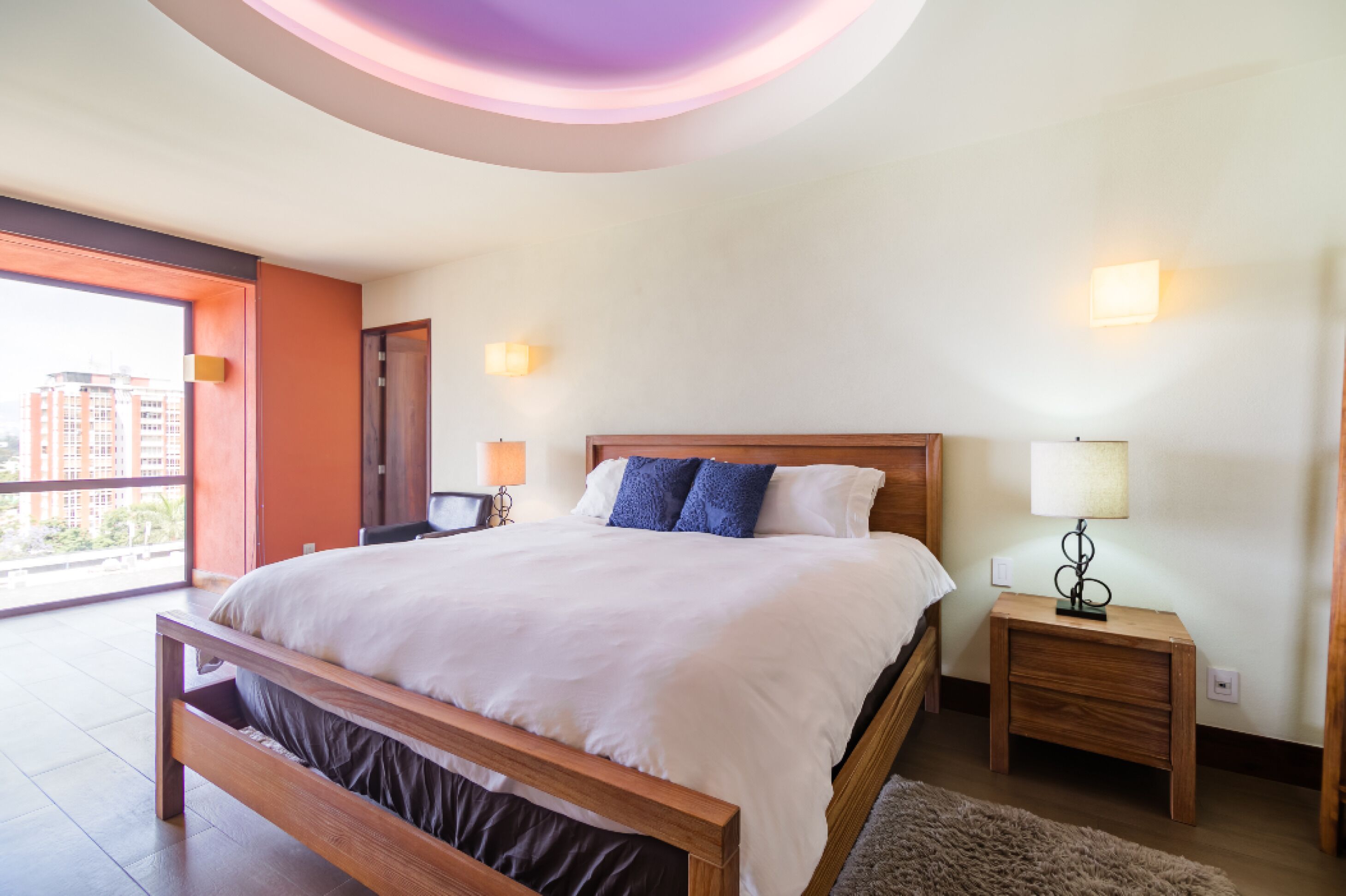 A bedroom with a large bed, wooden headboard, bedside tables with lamps, and a view of buildings through a large window.