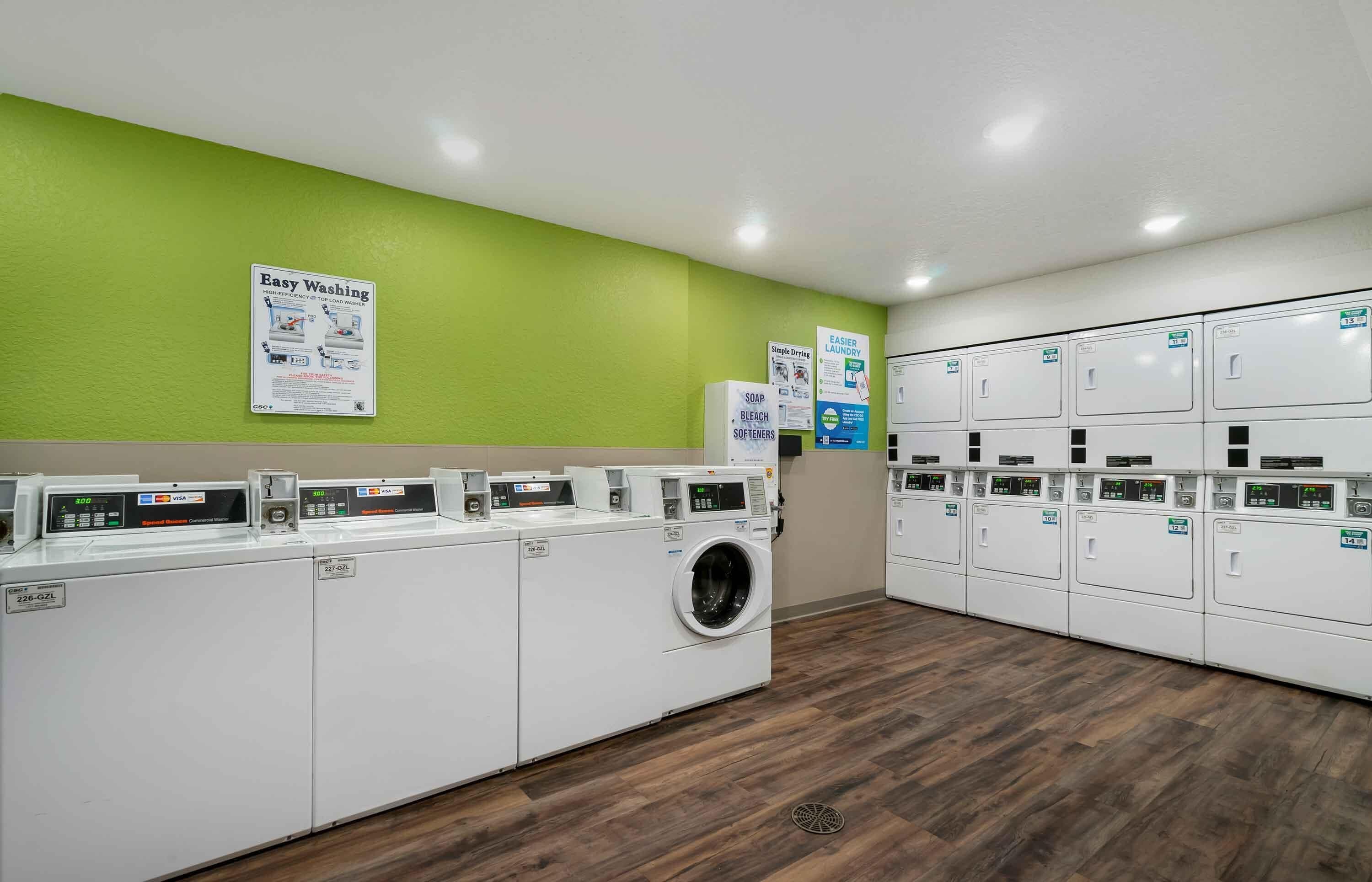 Un cuarto de lavandería con varias lavadoras y secadoras, una pared verde y carteles informativos.