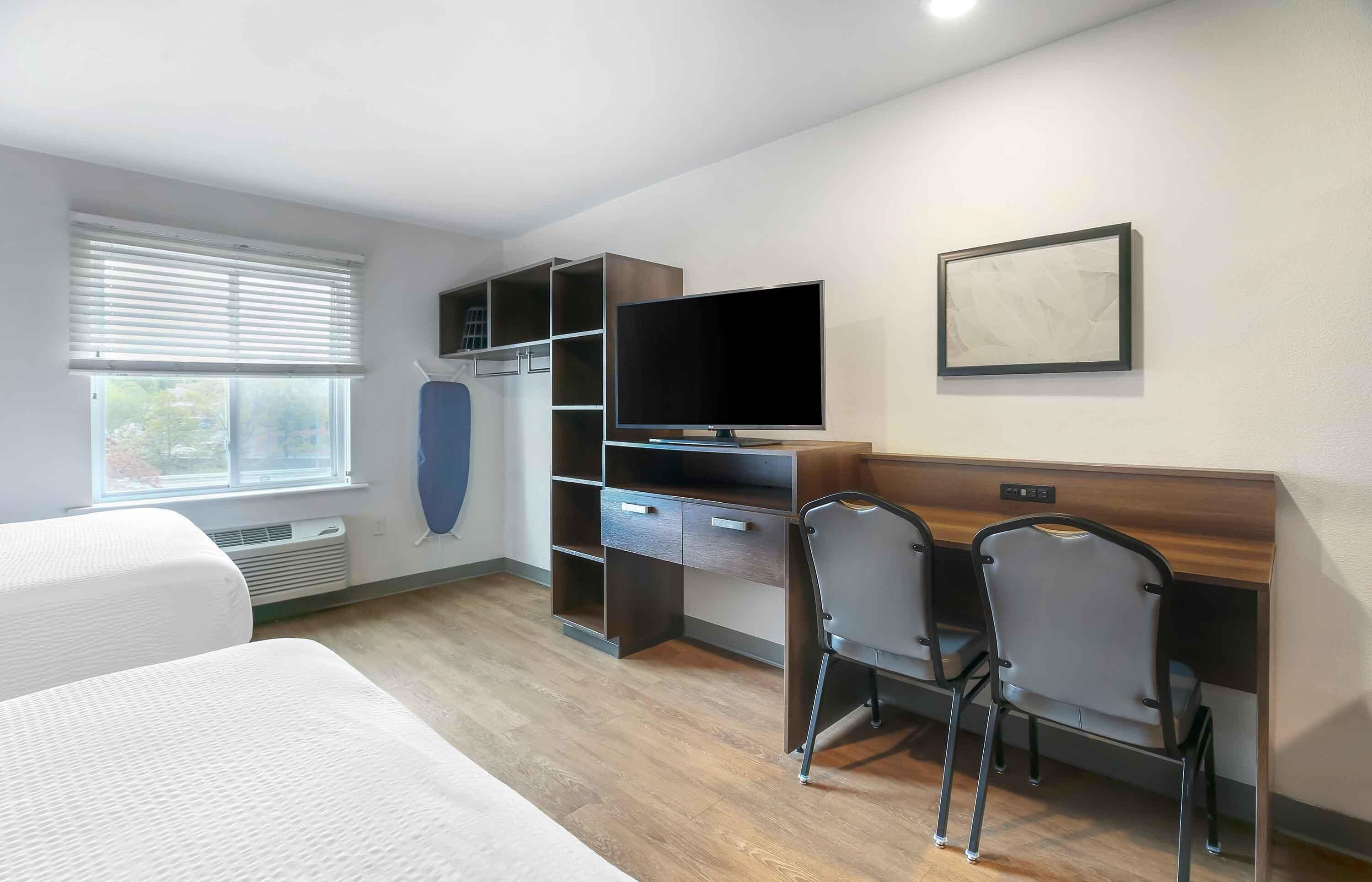 Habitación de hotel con cama, escritorio, computadora, televisor y ventana con persianas.