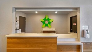 A reception desk with a wooden counter, a green abstract wall decoration, a door with a 'FACP' sign, and a 'CHECK-IN' and 'CHECK-OUT' sign on the wall. - Extended Stay America Select Suites Tampa Brandon East (Tampa)