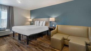 A hotel room with a large bed, a beige sofa, a nightstand with lamps, and a window with curtains. - Extended Stay America Select Suites Tampa Brandon East (Tampa)