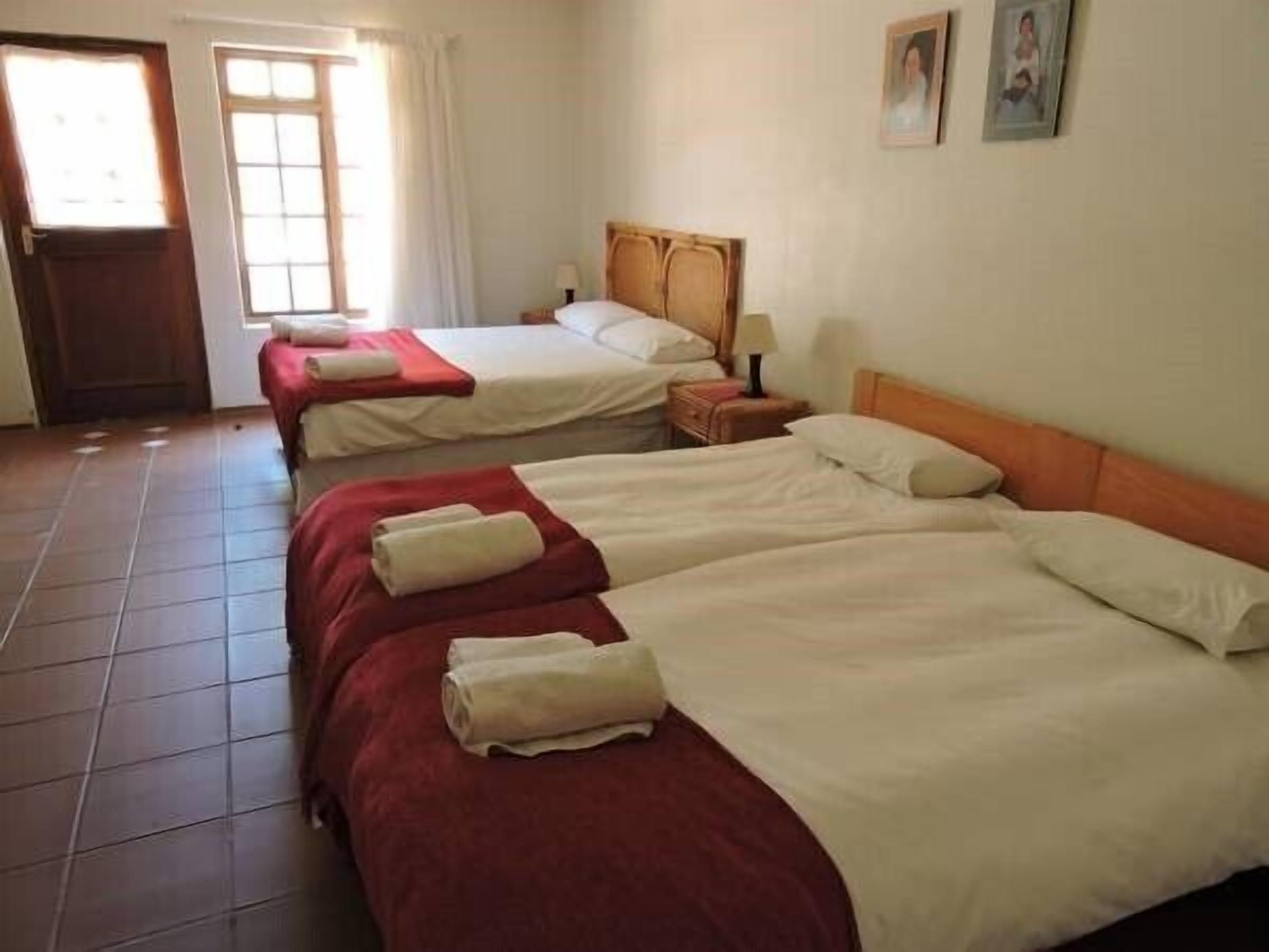 A hotel room with three beds, each with a red blanket and white sheets. There is a window with curtains, a door, and a small table with a lamp.