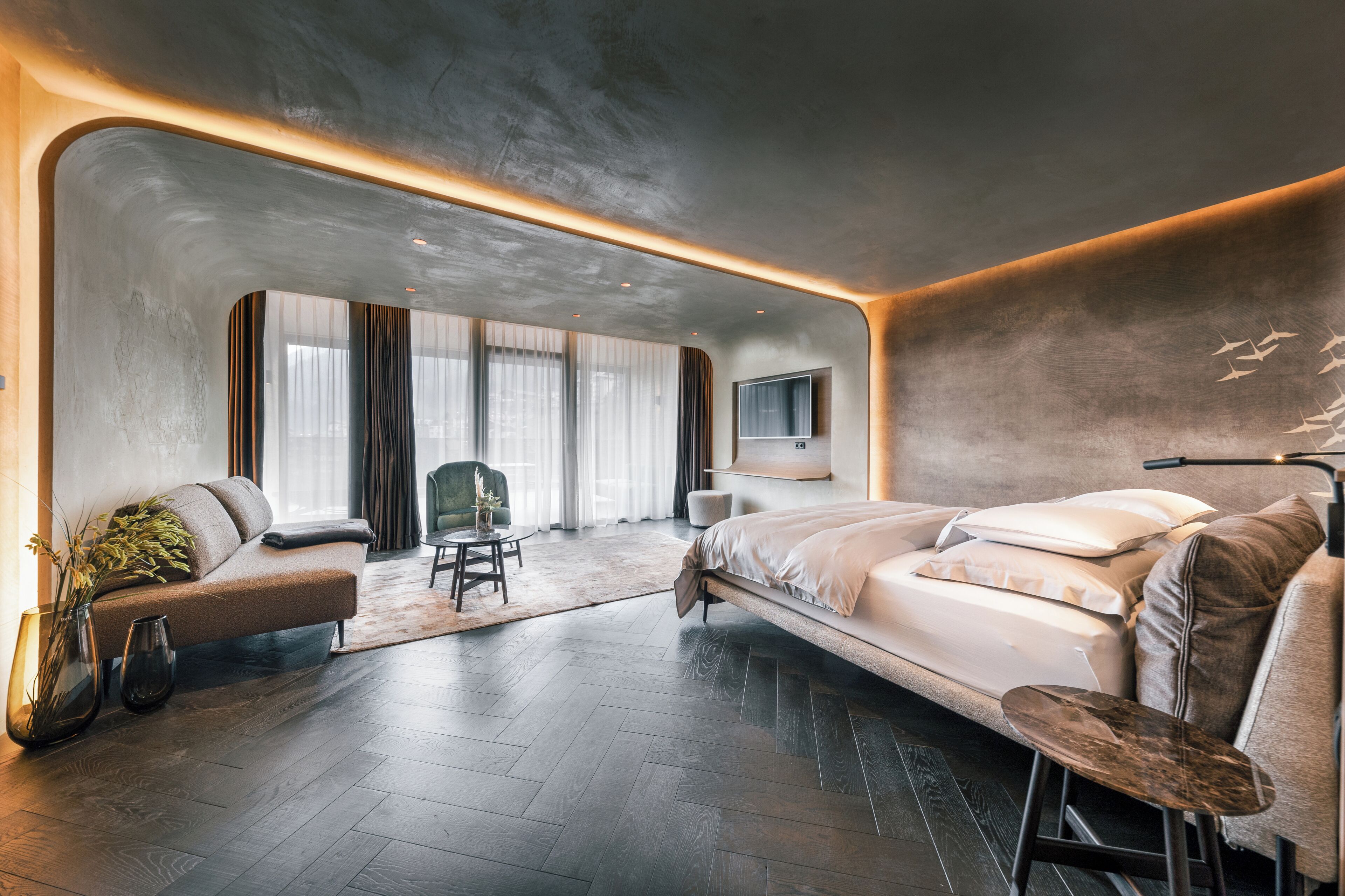 A modern hotel room with a large bed, a seating area with a chair, a small table, and a large window with curtains.