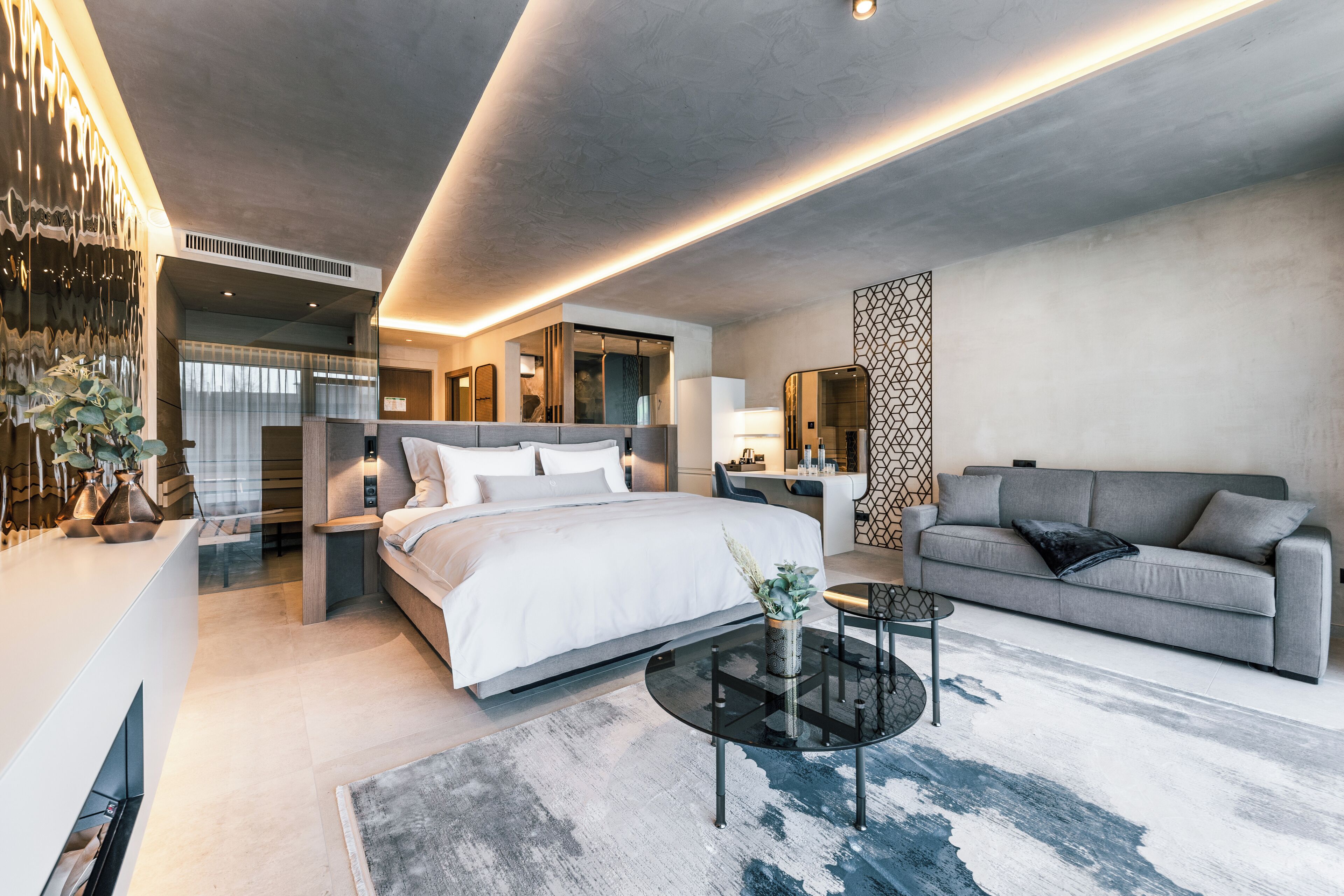 Una habitación de hotel moderna con una cama grande, un sofá gris, una mesa de centro de cristal y un panel decorativo en la pared.