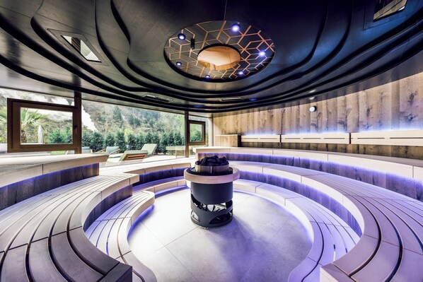 Un área de estar moderna y circular con una chimenea central, rodeada por un muro de madera y grandes ventanales que ofrecen vistas a la vegetación.