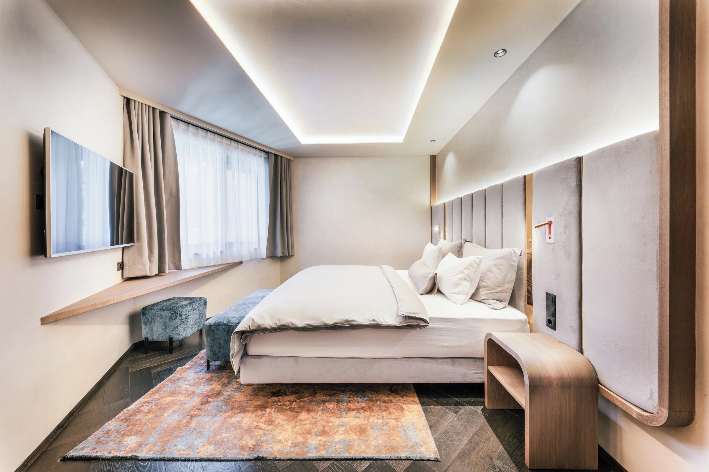 A modern hotel room with a bed, a seating area, a large window with curtains, and a mounted television.