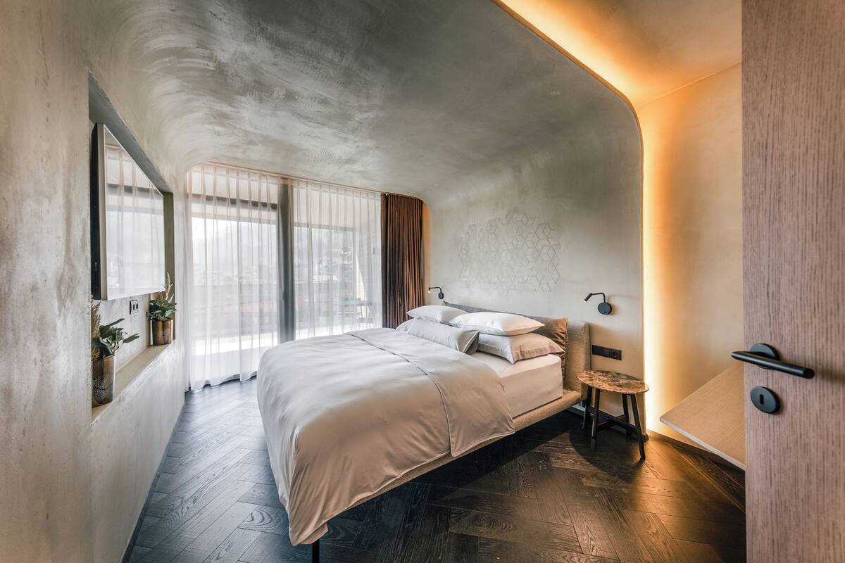 Une chambre moderne avec un grand lit, une petite table et une chaise. On y trouve des fenêtres avec des rideaux, un luminaire fixé au mur et une plante posée sur une étagère.