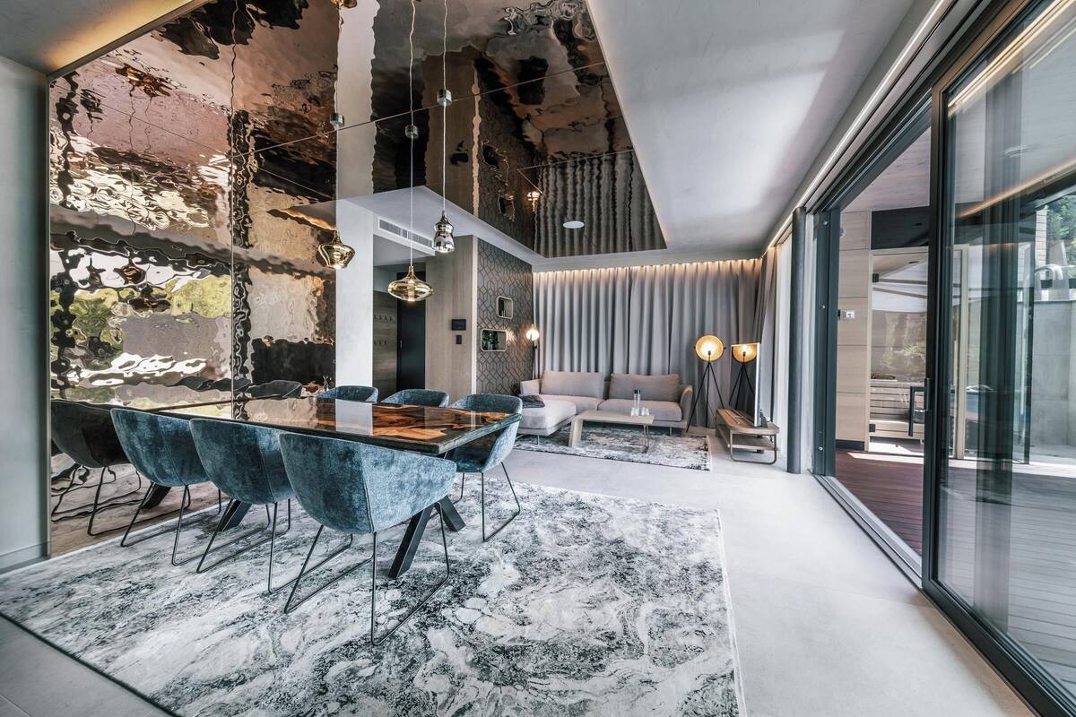 Un intérieur moderne comprenant un coin repas avec une table en verre et des chaises bleues, un coin salon avec un canapé et une table basse, et une grande fenêtre offrant une vue sur l’extérieur.