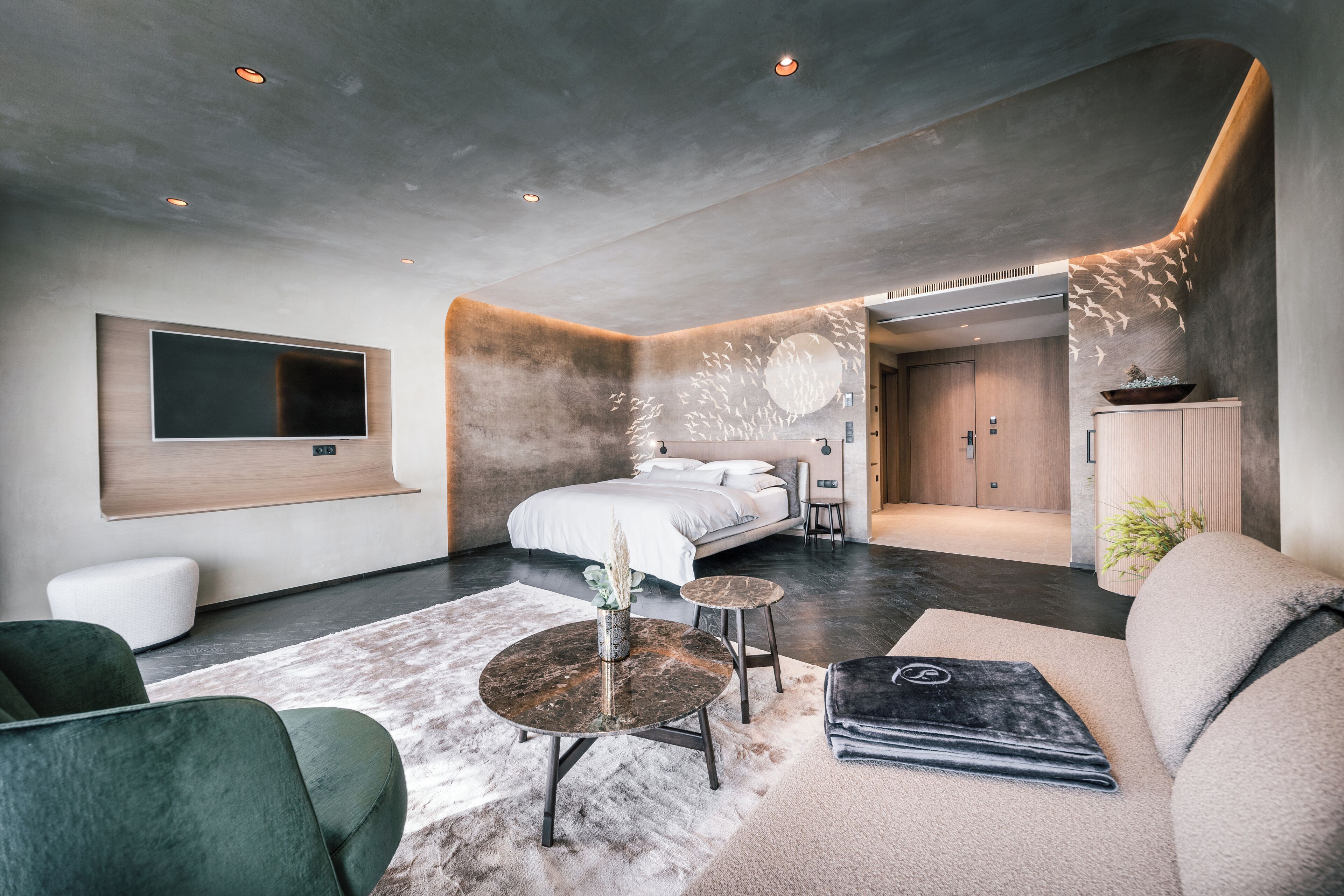 Une chambre d’hôtel moderne dotée d’un grand lit, d’un téléviseur fixé au mur, d’un coin salon avec un canapé et des fauteuils, et d’une petite table sur laquelle est posé un vase.