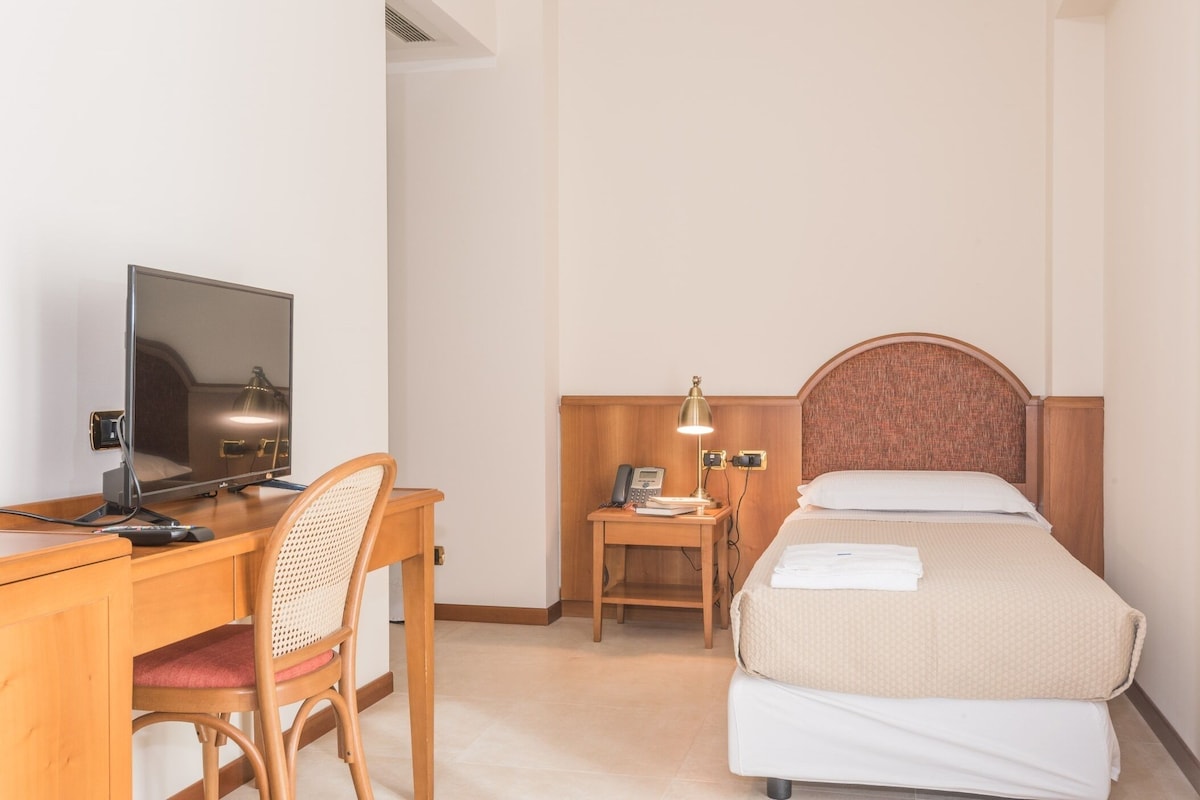 Habitación de hotel con cama, escritorio con computadora, silla, mesita de noche con lámpara y teléfono.