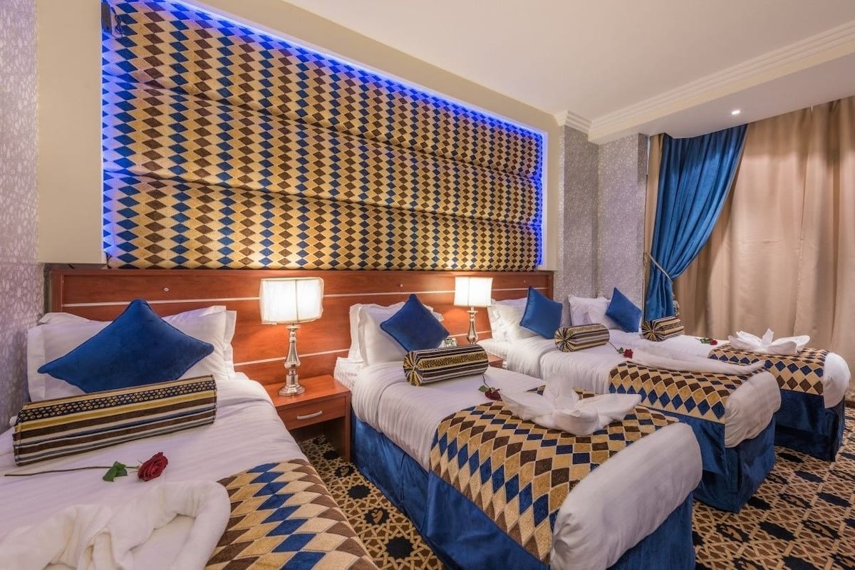 Ein Hotelzimmer mit vier Betten, jedes mit einer gemusterten Tagesdecke und blauen Kissen. An den Nachttischen stehen Lampen und über den Betten ist ein dekoratives Wandpaneel angebracht.