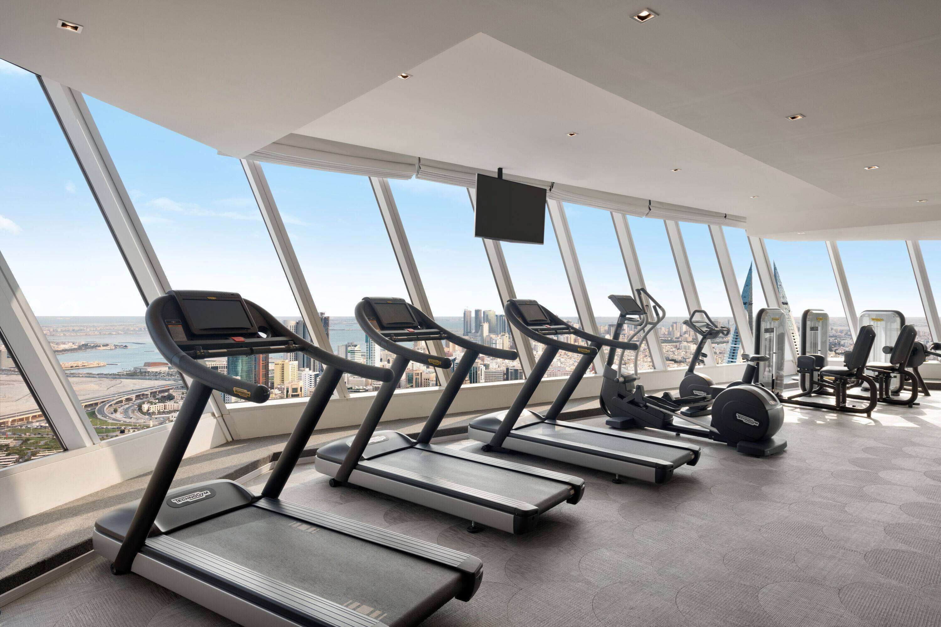 A gym with treadmills and exercise equipment, large windows with a city view.
