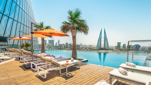 A rooftop pool area with lounge chairs and umbrellas, a palm tree, and a city skyline in the background. - Wyndham Grand Manama (Manama)