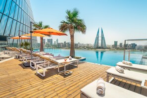 A rooftop pool area with lounge chairs and umbrellas, a palm tree, and a city skyline in the background. - Wyndham Grand Manama (Manama)