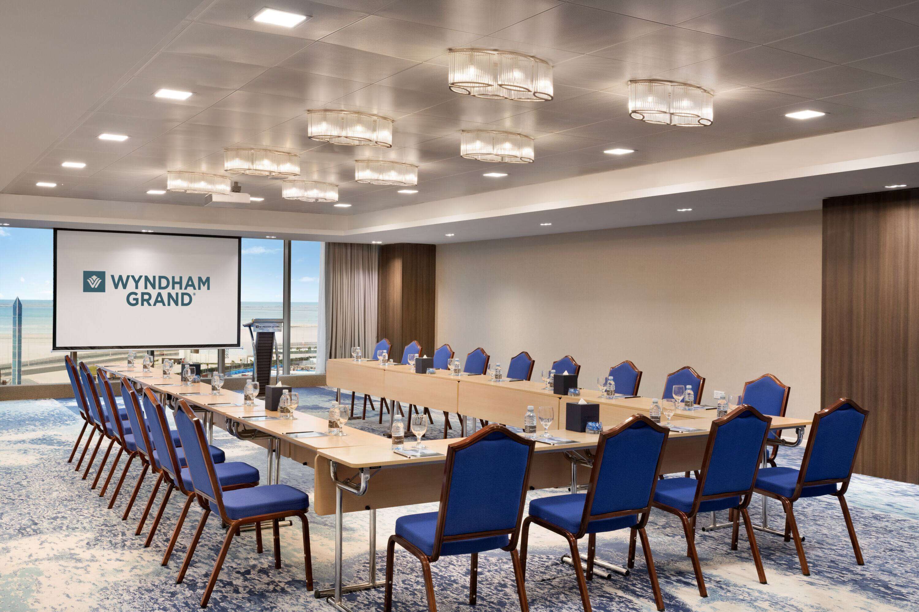 Un salón de conferencias con una mesa larga, sillas azules y una pantalla mostrando 'WYNDHAM GRAND'.