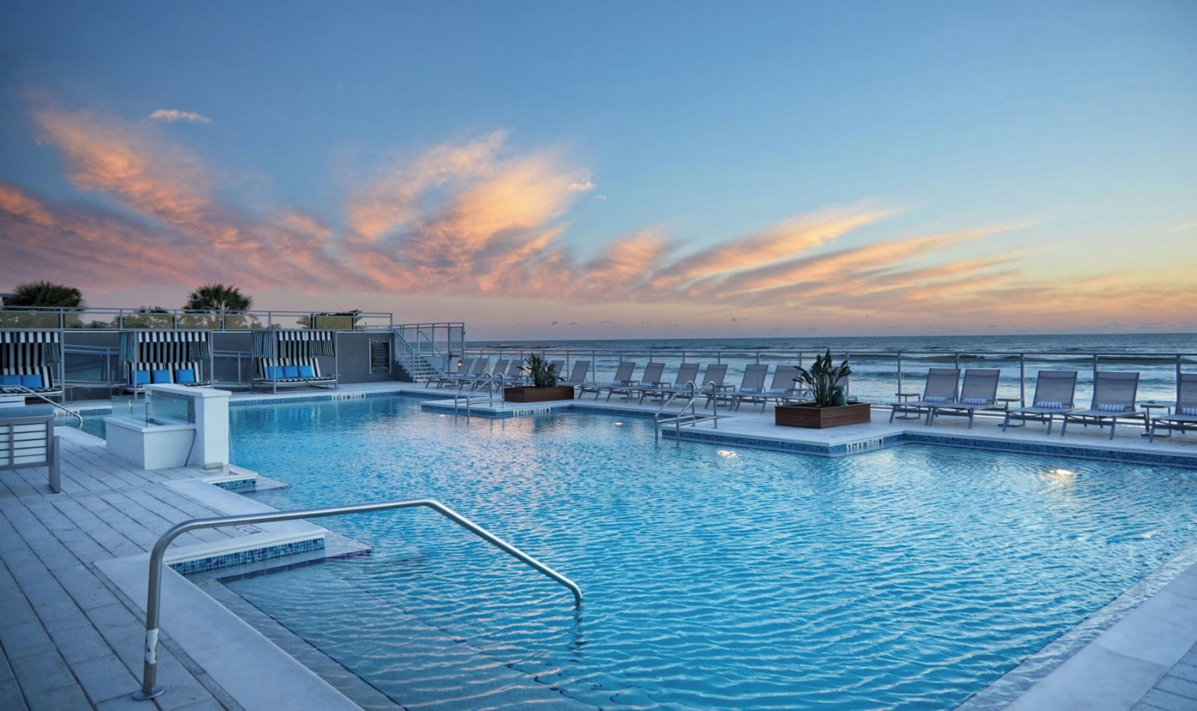 A pool area with multiple lounge chairs and a view of the ocean at sunset.