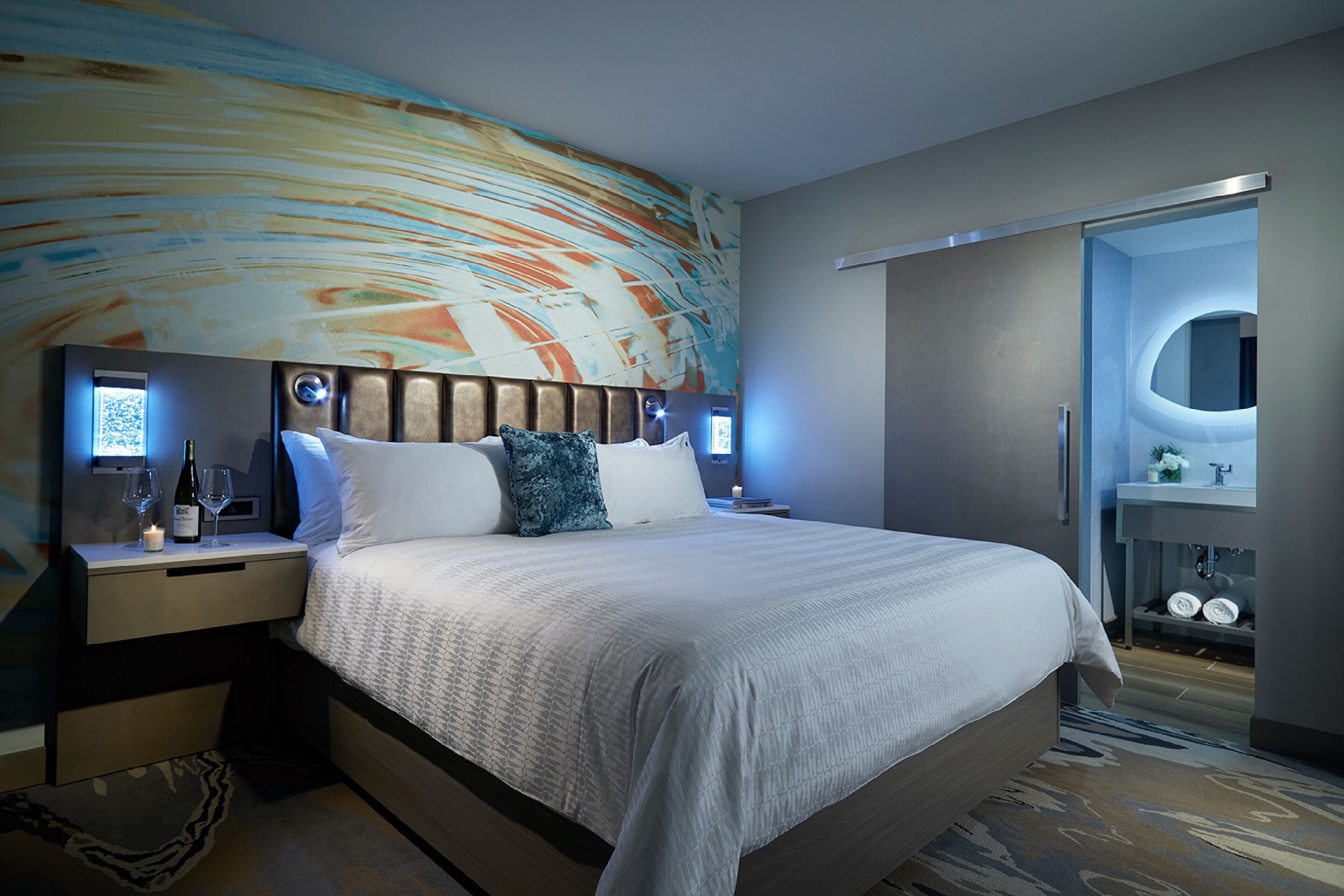 Quarto de hotel moderno com uma cama grande, uma mesa de cabeceira com uma garrafa de vinho e copos, uma cabeceira com iluminação circular e um banheiro com espelho e pia.