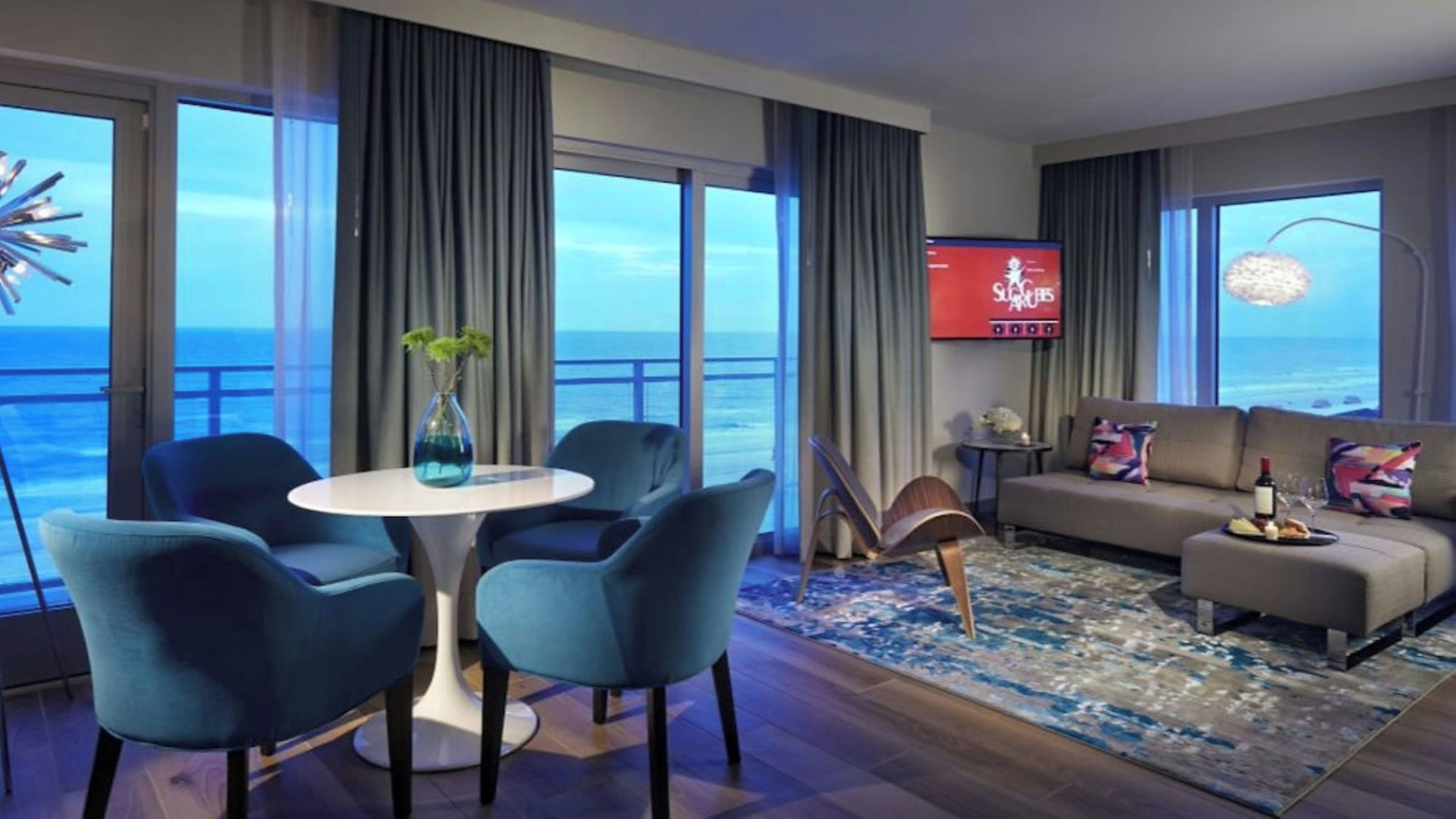 Une chambre d’hôtel moderne dotée d’une grande fenêtre donnant sur l’océan, d’une table à manger ronde avec des chaises, d’un canapé agrémenté de coussins décoratifs et d’un téléviseur à écran plat fixé au mur.